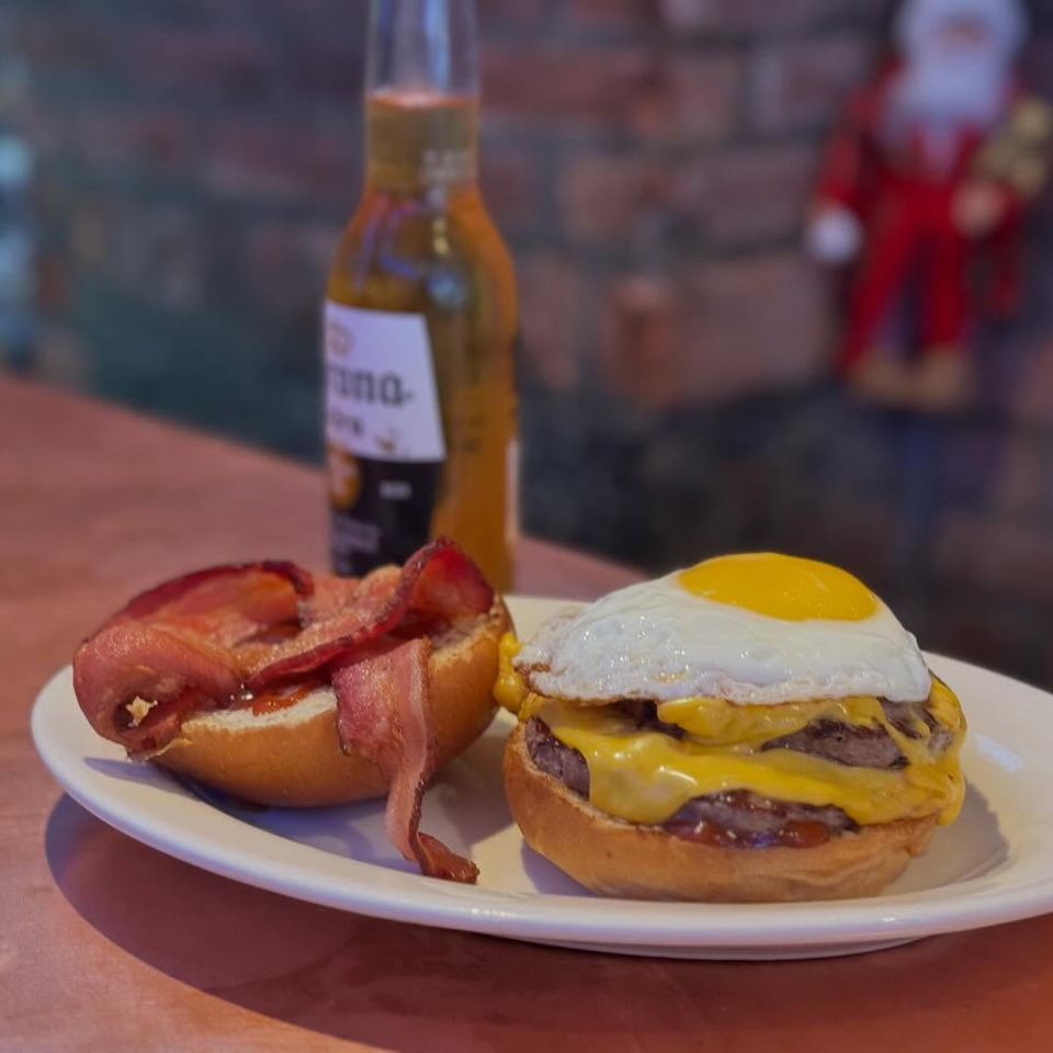 A double cheeseburger with a fried egg and bacon on a bun, next to a beer bottle.