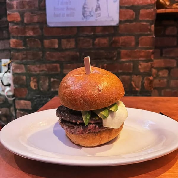 A double patty burger with avocado and white sauce on a white plate, against a brick wall.