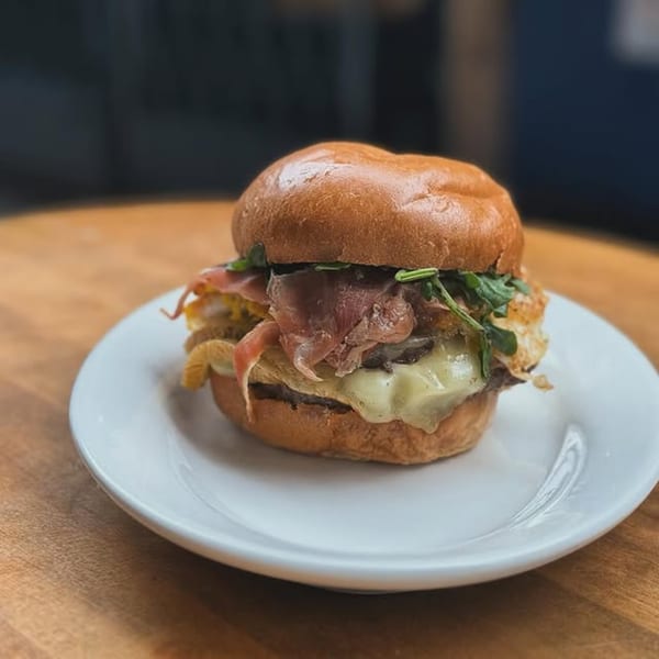 A gourmet burger with prosciutto, melted cheese, and arugula on a white plate.