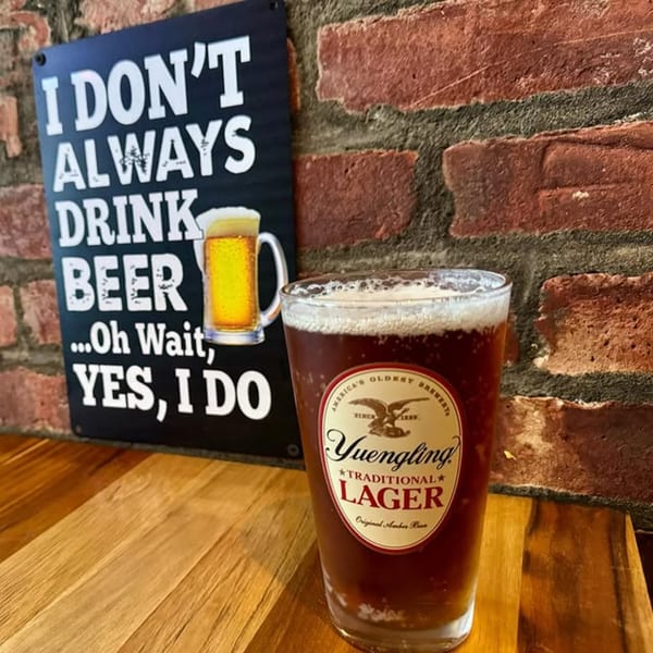 A glass of Yuengling Traditional Lager on a wooden table next to a humorous beer sign.