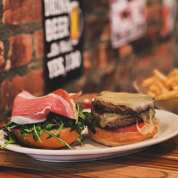 Two gourmet burgers, one with prosciutto and arugula, the other a double cheeseburger, on a white plate.